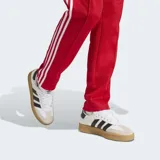 Pantalón deportivo Adidas de corte ajustado, color rojo con las icónicas tres tiras blancas laterales y logo del trébol bordado en la parte superior. Confeccionado en tejido de piqué con cintura elástica y cordón de ajuste.
