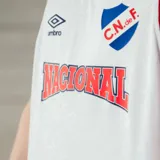 Camiseta de básquet Umbro del Club Nacional de Football. Diseño blanco con detalles en azul y rojo en cuello y sisas. Incluye logo de Umbro, escudo del club y sponsor Antel en el frente.