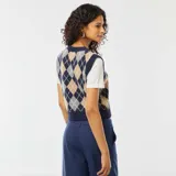 Chaleco de punto sin mangas, con cuello en V, de estilo argyle en tonos azul marino, beige, gris y blanco. Se muestra sobre una camiseta blanca de manga corta y un pantalón azul marino.
