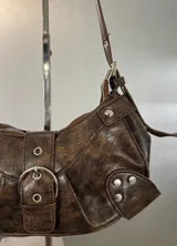 Cartera tipo hobo de cuero sintético con efecto desgastado, color marrón. Presenta una hebilla central decorativa, detalles de tachas metálicas en los laterales y correa ajustable.