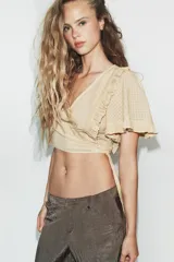 Top crop de escote en pico y manga corta con volados. Presenta un diseño cruzado que se ajusta mediante una lazada lateral y cuenta con una textura de cuadrícula sutil en toda la prenda.