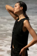 Chaleco de denim celeste sin mangas, confeccionado en algodón. Presenta un cierre frontal con botones forrados en la misma tela y un diseño de bajo redondeado con una abertura central.