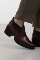 Botín asimétrico de estilo Chelsea color bordó, confeccionado en cuero vacuno, con paneles elásticos laterales negros, puntera cuadrada, suela de plataforma y tacón de bloque cuadrado.