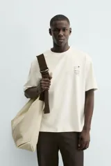 Camiseta de corte relaxed fit, color blanco roto, con cuello redondo y manga corta. Presenta un pequeño estampado en el pecho con un dibujo lineal y texto.