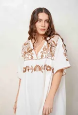 Vestido corto de corte amplio, color blanco, con escote en V y mangas cortas holgadas. Presenta detallados bordados en hilo marrón en la zona del pecho, los hombros y las mangas.