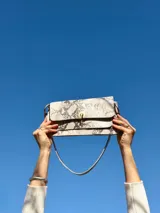 Cartera tipo mini bag con diseño de estampado de serpiente en tonos beige y marrón. Presenta una solapa frontal con cierre metálico dorado y una correa ajustable para llevar al hombro.