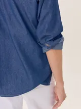Camisa de algodón color gris claro, con cuello clásico y detalle de volados en la cartera frontal. Presenta mangas largas con puños abotonados y un corte recto.