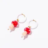 Aros tipo argolla con baño de oro, de color blanco, de los cuales cuelgan dijes de vidrio con forma de hongo rojo con puntos blancos y tallo color piel, acompañados de una pequeña cuenta blanca.