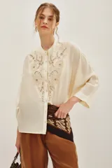 Camisa de viscosa color crema, con cuello mao y cierre frontal de botones. Presenta un delicado bordado floral en el frente y mangas tres cuartos con puños rectos.