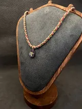 Collar de diseño artesanal compuesto por cordones trenzados multicolor en tonos verdes, rosados y rojizos. Presenta un dije de piedra ovalada con una cruz dorada aplicada en el centro.