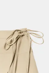 Falda mini de tiro medio en color beige, con diseño cruzado y bajo asimétrico. Presenta un cierre lateral con lazada ajustable.