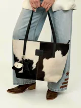 Cartera tote de cuero con pelo vacuno en tonos blanco y negro, con asas largas de hombro, parche frontal con logo y charms colgantes en forma de estrella y mini monedero.