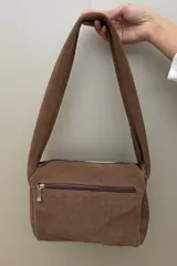 Cartera de hombro confeccionada en gamuza color habano, con diseño de correa ancha decorada con tachas metálicas circulares. Presenta cierre superior y placa metálica con logo en el frente.