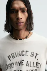 Camiseta de manga corta y cuello redondo, color blanco roto con efecto lavado y desgastado. Presenta un estampado frontal en negro con texto de estilo universitario que incluye "PRINCE ST.", "GROOVE ALLEY", "SOHO, NYC" y el número "95".