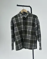 Camisa de manga larga con diseño de cuadros escoceses en tonos verde oliva, blanco y negro. Presenta cuello clásico, cierre frontal con botones y corte holgado.