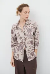 Camisa de manga larga con cuello clásico y cierre frontal de botones. Presenta un estampado floral en tonos marrones sobre una base color beige, confeccionada en un tejido con textura similar al gamuzado.