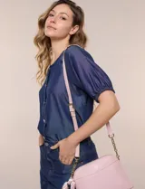 Cartera bandolera estructurada de eco cuero rosado con textura graneada, silueta curva y cierre superior. Posee correa larga regulable con detalles de cadena dorada, bolsillo frontal con logo metálico, pañuelo estampado a contratono anudado en un lateral y base rígida con apoyos metálicos.