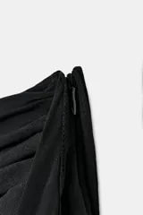 Falda pantalón de tiro bajo en color negro, con cintura drapeada y detalle de tira lateral colgante. Presenta un diseño con volantes en el bajo y cierre de cremallera metálica.