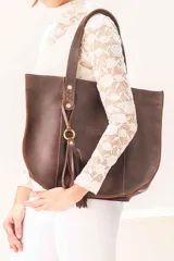 Bolso tipo shopper de cuero vacuno color chocolate. Presenta una silueta amplia y estructurada, con asas largas para llevar al hombro, detalles de trenzado artesanal en la unión de las asas y un colgante decorativo con flecos y argolla metálica. Incluye el logo de la marca grabado en el frente.