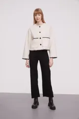 Chaqueta corta de paño color marfil, con cuello redondo, cierre frontal de botones negros y dos bolsillos frontales. Presenta detalles de costuras decorativas en contraste negro en los bordes de los bolsillos, puños y ruedo.