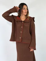 Saco marrón de punto calado con cuello bobo amplio con volados y detalles bordados en hilo rojo. Presenta cierre frontal de botones, mangas largas con puños acampanados y costuras decorativas en contraste.