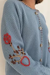 Saco de punto color beige, confeccionado en algodón, con cuello redondo, mangas largas y cierre frontal con botones. Presenta detalles bordados con diseño floral en las mangas y ribetes con rayas azules y blancas en la botonera y el cuello.