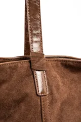 Bolso tipo tote bag color marrón, confeccionado en gamuza vegana con detalles de tiras y asas de hombro en cuero sintético brillante. Incluye una placa metálica pequeña con el logo de la marca en el frente.