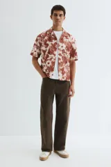 Camisa de manga corta con cuello abierto, tapeta de botones y bajo recto. Presenta un diseño estampado con motivos florales en tonos tierra y blanco.