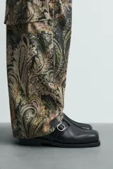 Zueco destalonado de cuero negro con hebilla metálica ajustable en el empeine, plantilla de cuero y suela plana con pequeño tacón.