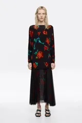 Vestido midi de corte ajustado con falda superpuesta de tul, escote barco y mangas largas tipo murciélago. Presenta un diseño estampado con motivos florales en tonos rojos y verdes sobre fondo negro, con detalles fruncidos en la cintura.