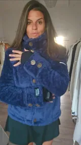 Campera corta de textura afelpada en color azul, con cuello alto y cierre frontal con botones dispuestos en diagonal. Presenta puños con aberturas para los pulgares y detalles de etiquetas visibles en las mangas.