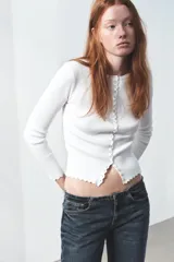 Chaqueta de punto acanalado en color blanco, con cuello redondo, manga larga y cierre frontal con botones nacarados. Presenta terminaciones con ondas en el ruedo y los puños.