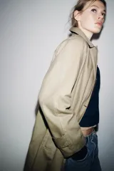Trench coat de corte largo y diseño minimalista, confeccionado en tejido encerado color beige. Presenta cuello solapa, cierre frontal con botones oscuros y bolsillos laterales integrados.
