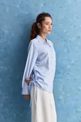 Camisa celeste de calce relajado, con cuello camisero, bolsillo aplicado en el pecho y mangas largas con puños abotonados. Cuenta con lazos ajustables en los laterales para fruncir y regular la silueta.