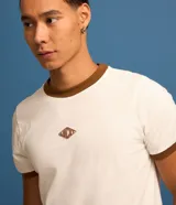Remera estilo baby tee de color blanco, confeccionada en algodón, con cuello redondo y ribetes en color marrón en cuello y puños. Cuenta con un pequeño tag gomosa con el logotipo "Lucky" en el centro del pecho.