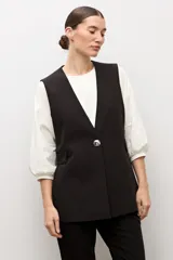 Chaleco largo sastrero de color negro, con escote en V profundo, cierre frontal con un botón metálico y bolsillos laterales con solapa.