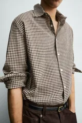 Camisa de corte relajado confeccionada en tejido de algodón con textura irregular y estampado de cuadros pequeños en tonos tierra. Presenta cuello solapa, cierre frontal de botones y mangas largas con puños abotonados.