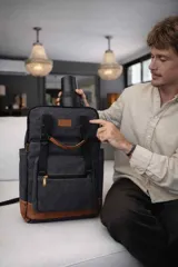 Mochila matera y porta notebook en tela canvas de color marrón, con diseño funcional que incluye bolsillo frontal con cierre, correas ajustables, manija superior y detalles de tiras de lona con hebillas metálicas.