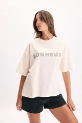 Remera manga corta de corte boxy en color crudo, con cuello redondo y estampa frontal con la palabra "BONHEUR" en tonos verdes y celestes.