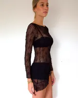 Vestido corto de encaje negro con diseño asimétrico en el ruedo. Presenta mangas largas y un corte ajustado, con un estilo que combina transparencia y texturas.