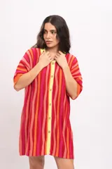 Vestido camisola corto de tejido fresco de algodón, con diseño a rayas verticales en tonos rojos, naranjas y fucsias. Cuenta con cuello polo y botonadura frontal en tono amarillo claro, y mangas cortas holgadas.