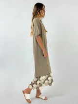 Vestido midi de tejido ligero color beige, con cuello en V, mangas cortas y ruedo decorado con aplicaciones de crochet en tonos tierra y rosado.