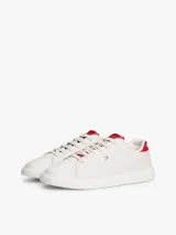 Zapatillas Tommy Hilfiger de diseño clásico y bajo, color blanco con detalles en rojo en la parte trasera del talón y en la lengüeta. Presentan cierre de cordones y el logo metálico distintivo de la marca en el lateral.