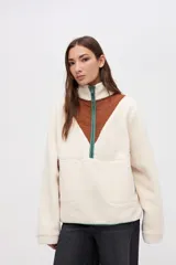 Buzo de sherpa color crema con diseño de medio cierre frontal. Presenta un panel triangular en el pecho de nylon color marrón y detalles de vivos en color verde en el cierre y bordes. Cuenta con cuello alto, mangas largas y bolsillos laterales.
