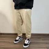 Pantalón tipo jogger de algodón color beige, con puños elásticos en los tobillos y estampa pequeña en el frente con el texto 'BUENA VIBRA EST. 2018'.