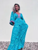 Túnica larga de estilo boho, color turquesa con estampado paisley en tonos azules y dorados. Presenta escote en V profundo con bordados decorativos en color fucsia y blanco, mangas amplias tipo kimono y ajuste en la cintura con cordón. El diseño incluye un ruedo con volado y terminaciones con borlas.