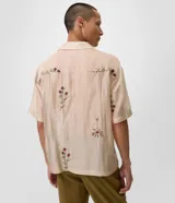 Camisa de lyocell color beige con corte boxy, cuello bowling y mangas cortas. Presenta un delicado bordado floral artesanal y cierre frontal con botones.