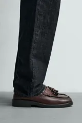 Mocasín de vestir color marrón oscuro, con parte superior lisa, detalle de antifaz con borlas en el empeine y horma redonda. Suela de goma a tono.