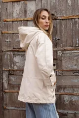 Parka liviana color beige con capucha, cierre frontal con botones, bolsillos delanteros con tapa y ajuste en la cintura. Presenta mangas con puños vueltos y detalle de botones.