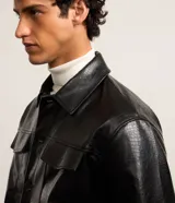 Campera trucker negra de cuero sintético con textura crocco, cuello inglés, cierre con botones, bolsillos con solapa en el pecho y puños abotonados.
